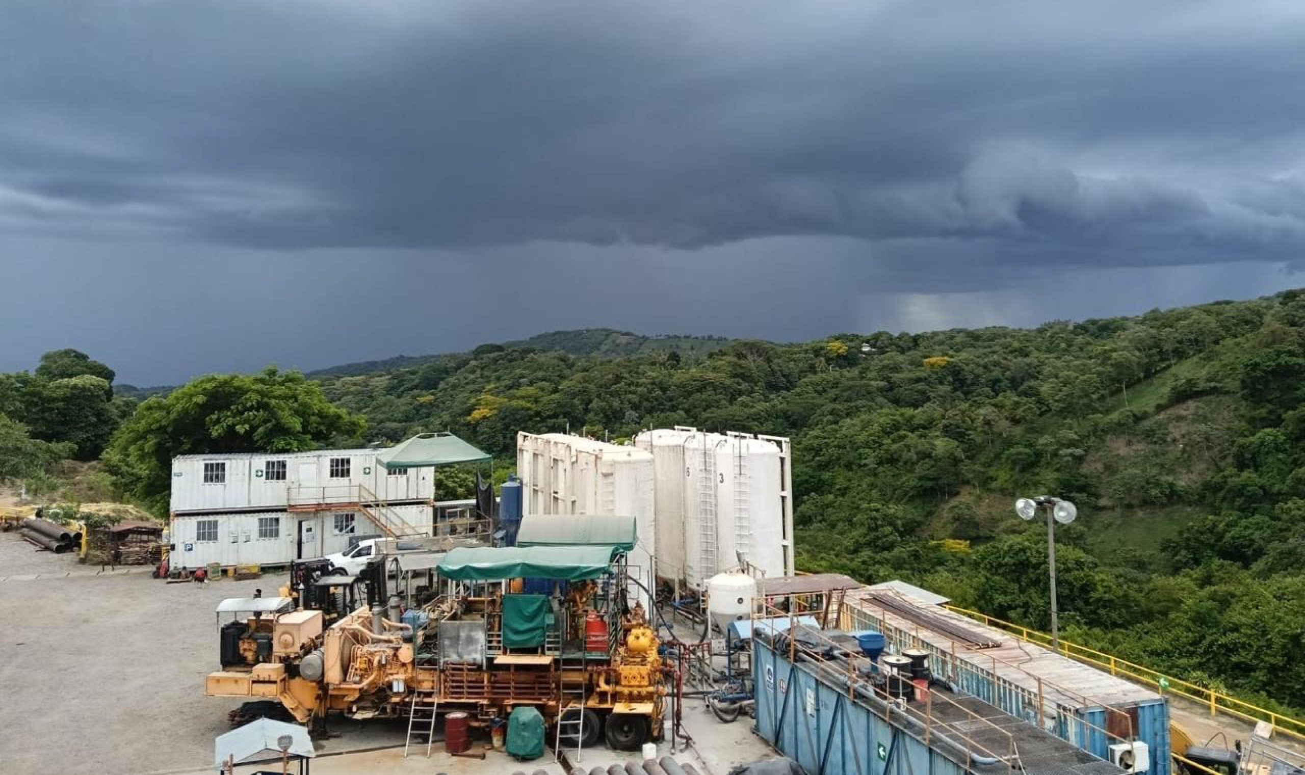 Geothermal well site with storage tanks below a green hillside under storm clouds